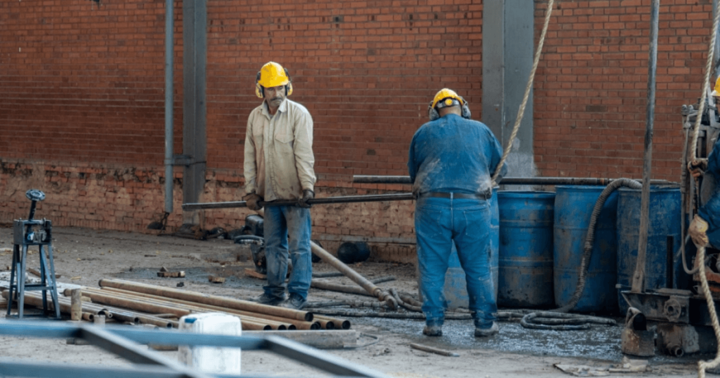 Obreros con cascos de seguridad trabajan en una obra de construcción, manipulando tuberías y equipos frente a una estructura de ladrillo.