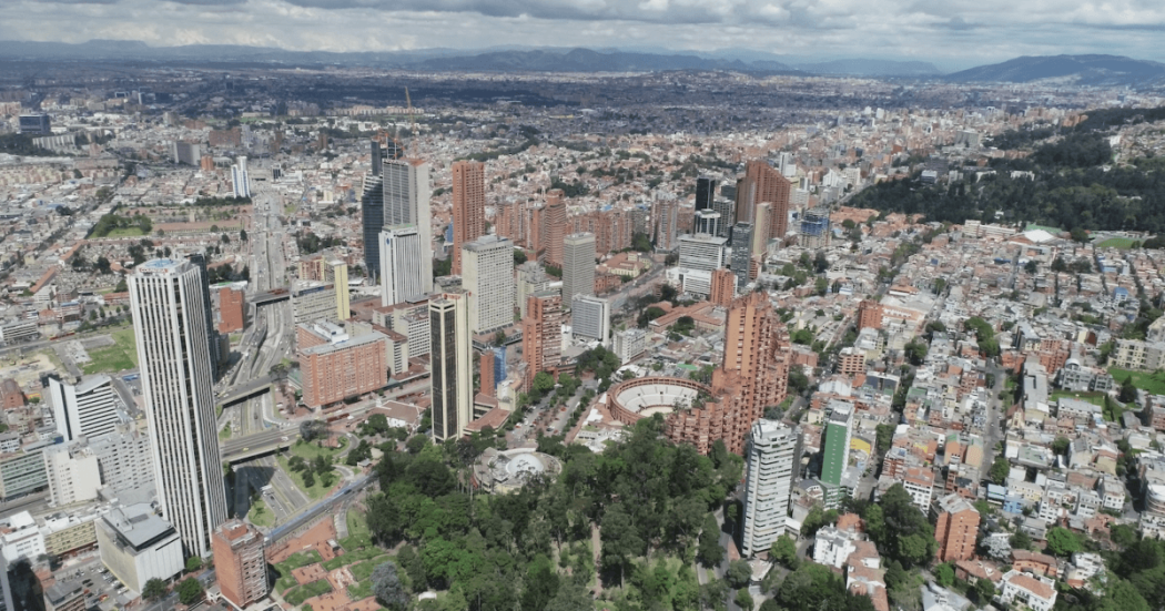 Vista aérea de un sector de Bogotá con edificios de gran altura, vías principales, zonas residenciales de alta densidad, áreas verdes y montañas al fondo bajo un cielo parcialmente nublado.