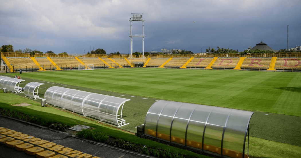 Estadio de Techo de Bogotá, con una cancha de fútbol de césped natural rodeada por graderías amarillas, bancas técnicas cubiertas y un cielo nublado al fondo.