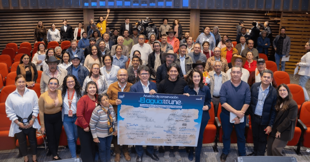 Grupo de personas reunidas en un auditorio con sillas rojas. En el centro de la imagen varias personas sostienen un tablero gigante con el texto: “Acuerdo de conservación. El agua que nos une”, junto a múltiples firmas