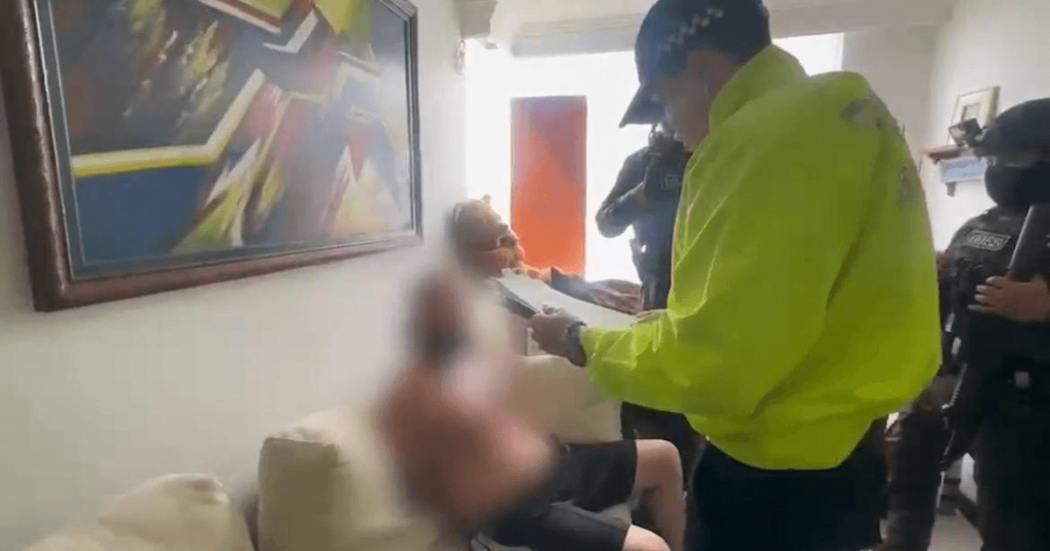 Persona con el rostro difuminado sentada en un sofá mientras otra persona con chaqueta reflectiva amarilla permanece de pie a su lado en una sala con cuadros en la pared