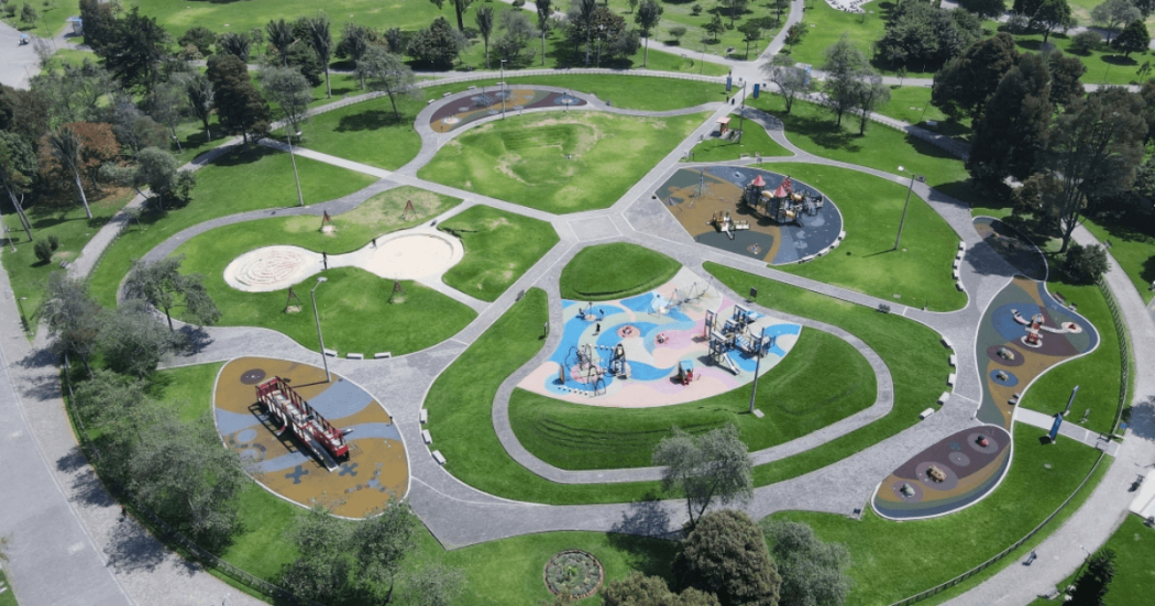 Vista aérea de un parque con amplias zonas verdes, senderos peatonales curvos y varias áreas de juegos infantiles distribuidas alrededor de una plazoleta central. Se observan estructuras de juego de diferentes tamaños, superficies de colores y árboles rodeando el espacio recreativo.