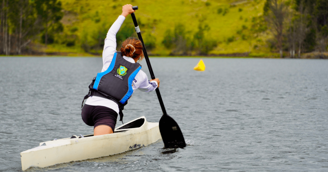 Persona practicando canotaje en un lago rodeado de vegetación, mientras impulsa una embarcación con un remo sobre el agua.