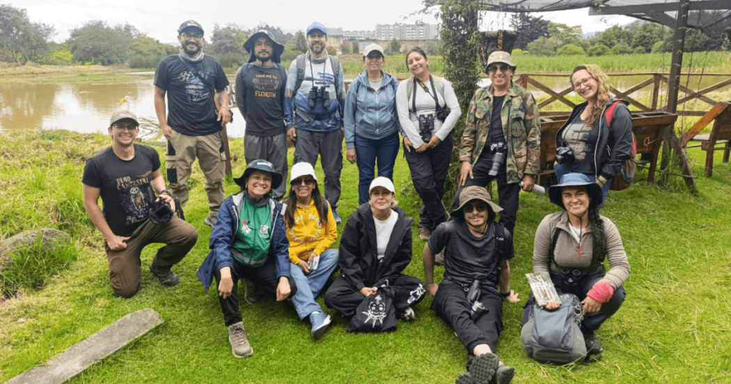 Grupo de 13 personas posa sobre una zona de césped del humedal Capellanía. Varias llevan binoculares, cámaras fotográficas y sombreros para observación de fauna.