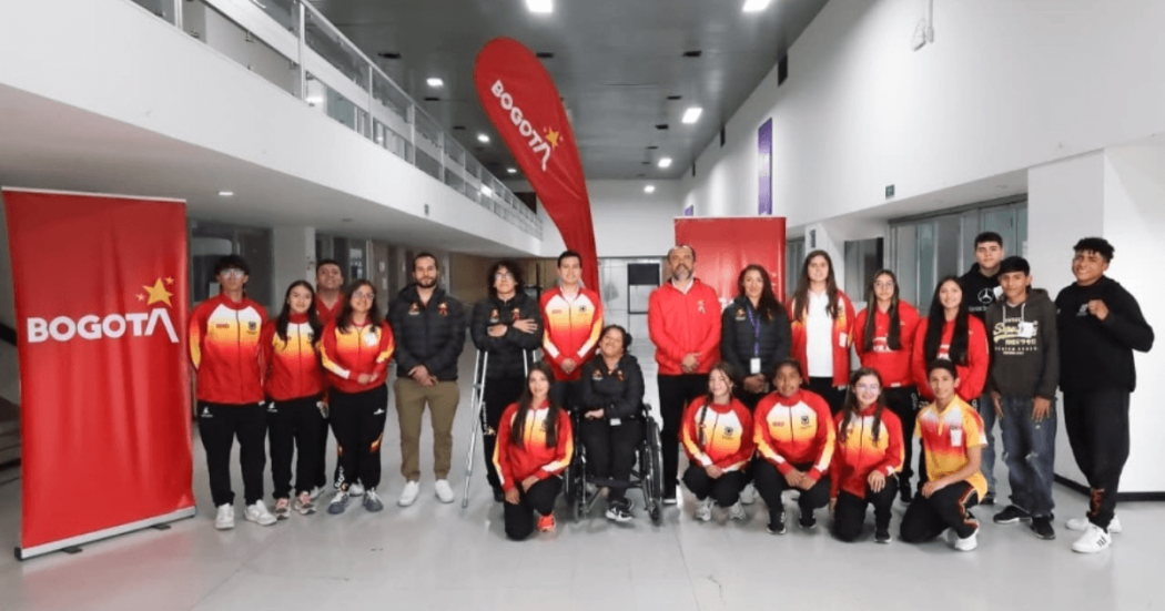 Grupo de deportistas del Equipo Bogotá, junto a personal de apoyo, posan para una fotografía en el hall interior de un edificio. Varias personas visten chaquetas deportivas en tonos rojo, amarillo y blanco.