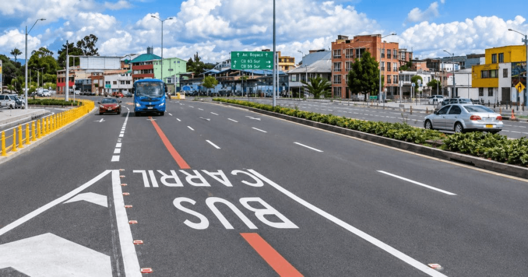 Carril preferencial para buses en la calle 63 de Bogotá, con un bus azul circulando por la vía, señalización vial pintada sobre el pavimento, separadores con vegetación y edificios al fondo.