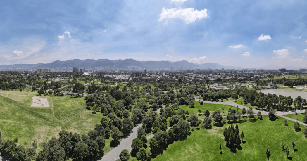 Vista panorámica aérea de un parque con extensas zonas verdes, senderos peatonales, grupos de árboles y un lago en un costado.