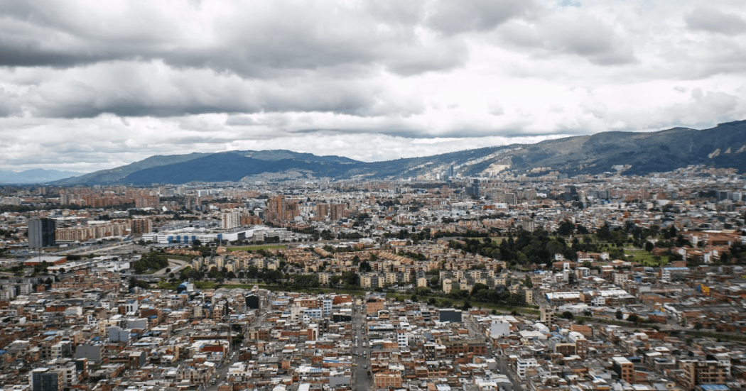 Vista panorámica de Bogotá con áreas residenciales y comerciales, edificios de diferentes alturas, vías urbanas y montañas al fondo bajo un cielo cubierto de nubes grises.