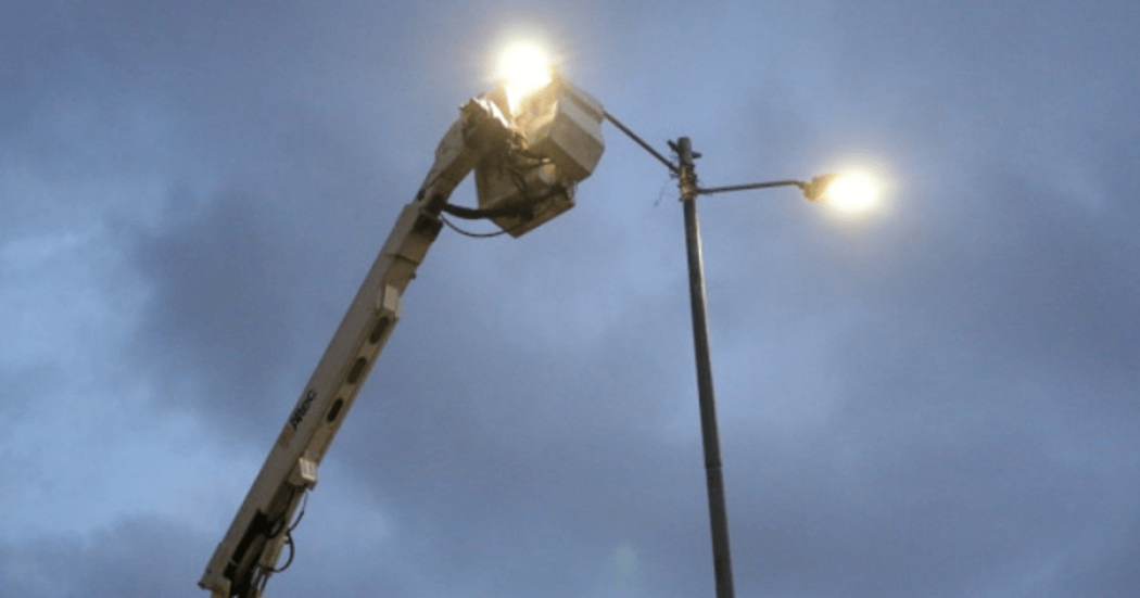 Poste de alumbrado público encendido mientras una plataforma elevadora realiza labores de mantenimiento en la parte superior, bajo un cielo nublado al atardecer.