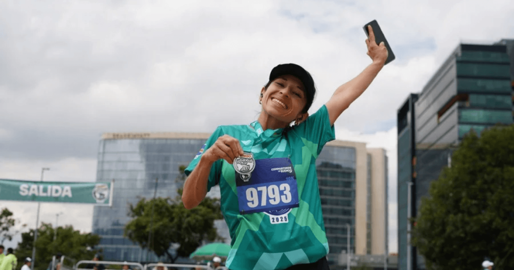 Participante de la Bimbo Global Race con camiseta verde y dorsal 9193 levanta un brazo mientras corre. Al fondo se observan edificaciones y el cielo despejado.