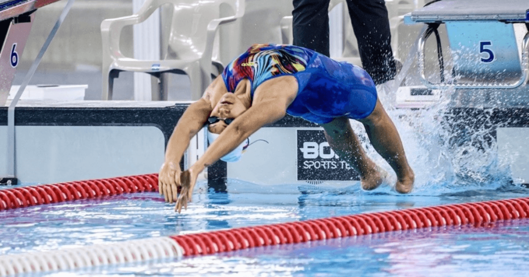 Nadador realiza la partida de espalda desde el borde de una piscina de competencia. La deportista viste traje de baño azul, gafas y gorro de natación, mientras se arquea hacia atrás para ingresar al agua.