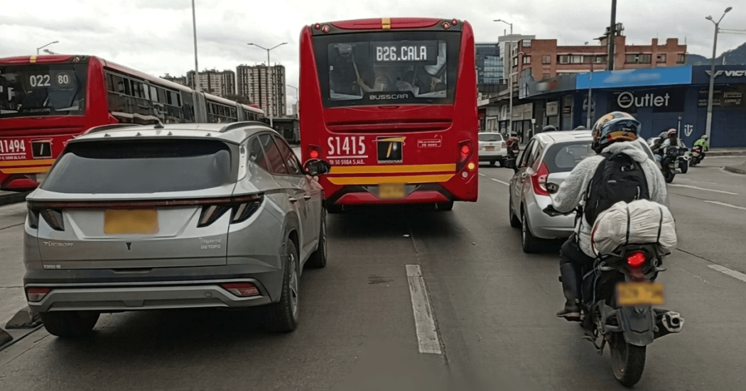 Bus articulado rojo de TransMilenio circula por una vía urbana junto a un automóvil gris y una motocicleta con carga en la parte trasera.