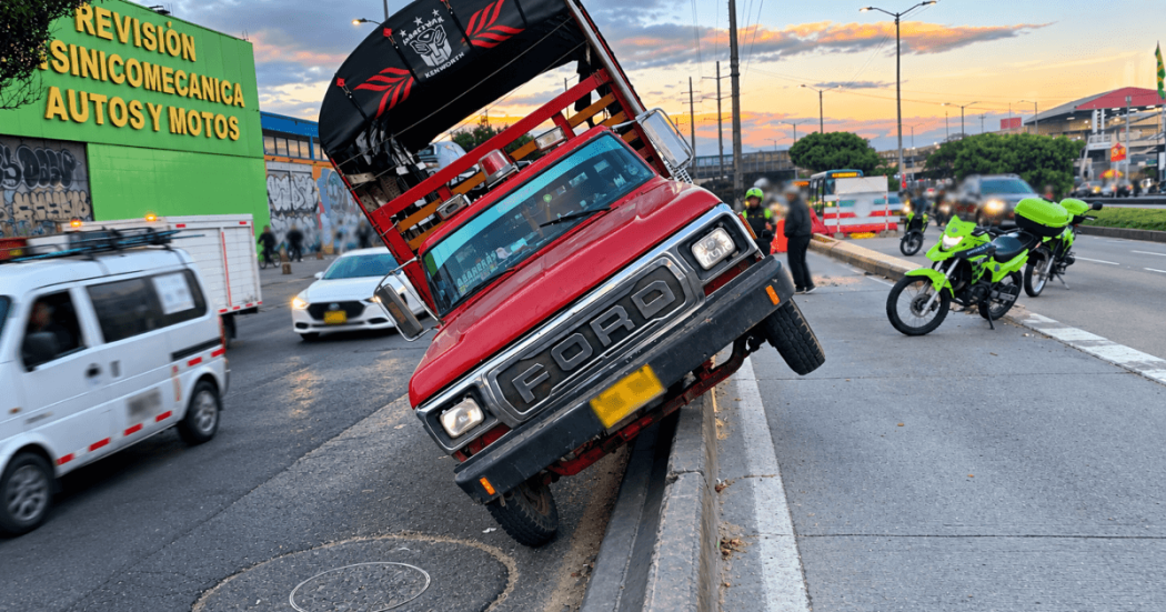 Camión rojo inclinado sobre el separador de una vía de Bogotá, con dos ruedas elevadas. A un costado hay motocicletas de tránsito, personal en la calzada y vehículos circulando en varios carriles.