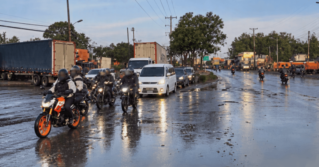 Motocicletas, vehículos particulares y camiones circulan por una vía mojada tras una lluvia, con reflejos sobre el pavimento y tránsito lento en ambos sentidos. Al fondo se observan árboles, postes de energía y señalización vial.