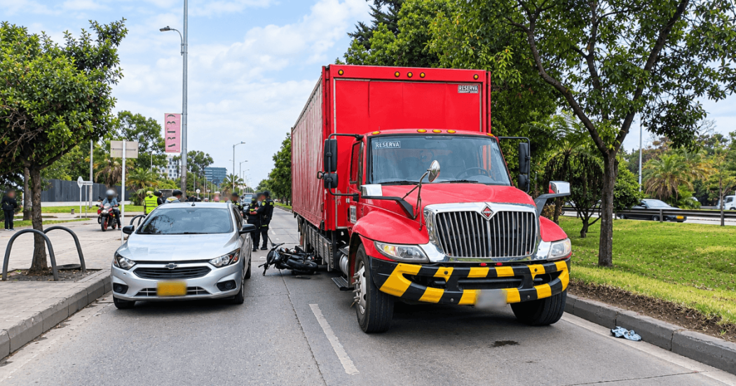 Vehículo de carga rojo y automóvil gris detenidos sobre una vía de Bogotá, junto a una motocicleta caída entre ambos. En el lugar hay presencia de agentes de tránsito y otras personas, con zonas verdes y árboles a los costados de la carretera.