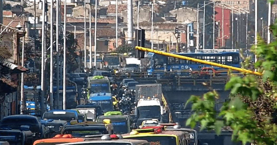 Panorámica de una vía con alta congestión vehicular en Bogotá. Se observan buses de TransMilenio, automóviles, motocicletas y vehículos de carga circulando en varios carriles, junto con agentes de tránsito en la calzada.