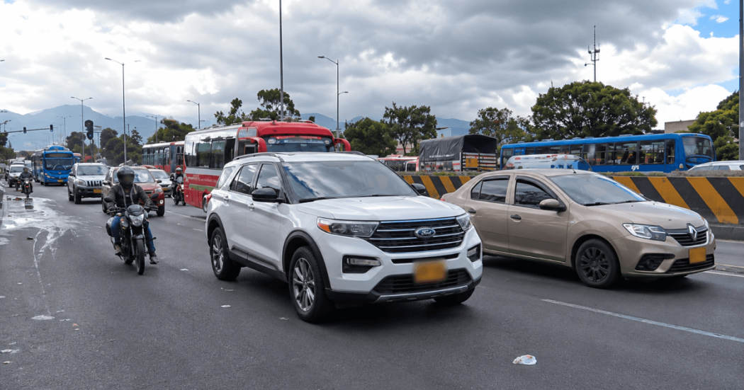 Vehículos particulares, motocicletas y buses circulan por una vía de varios carriles en Bogotá bajo un cielo nublado.