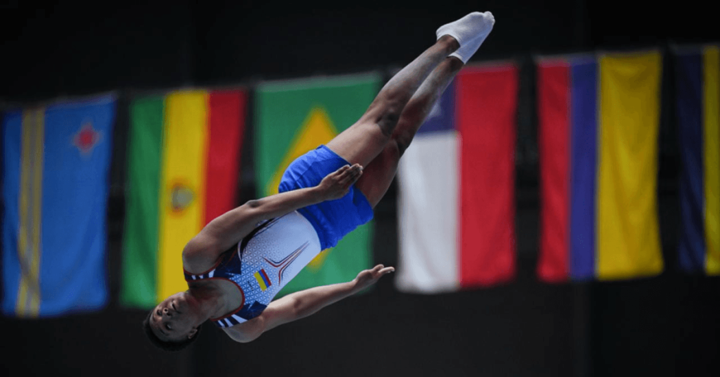 Deportista del Equipo Bogotá ejecuta un salto en competencia de atletismo, captado en el aire durante la prueba. Al fondo se observan banderas de distintos países participantes.