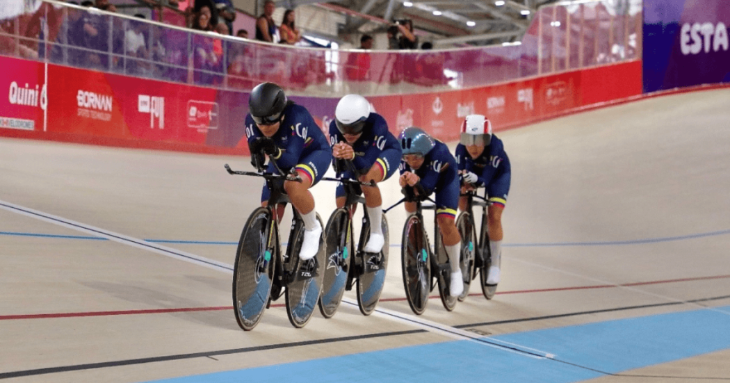 Cuatro ciclistas colombianas compiten en una pista de ciclismo de velódromo, avanzando en formación sobre sus bicicletas de pista mientras usan uniformes azul oscuro y cascos de protección.