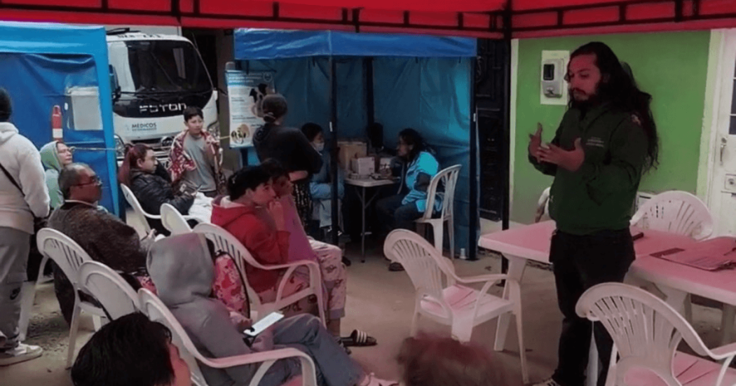 Personas sentadas en sillas plásticas asisten a una charla en un espacio cubierto, mientras una persona expone junto a una mesa.