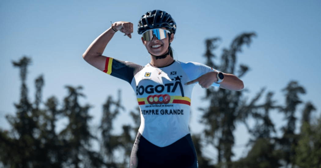 Gabriel Rueda, patinadora bogotana, posa con uniforme de la Liga de Patinaje de Bogotá y casco deportivo en una pista al aire libre, señalando el logo de Bogotá en la camiseta, con árboles desenfocados al fondo y cielo despejado.