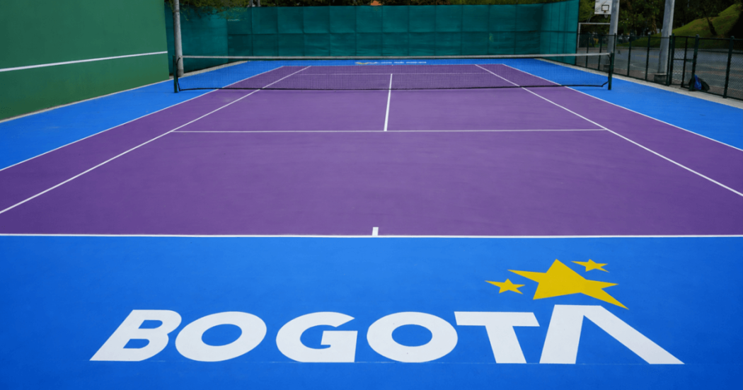 Cancha de tenis con superficie morada y zonas laterales azules, delimitada por líneas blancas y una red en el centro. En el primer plano se observa el logo de Bogotá en color blanco y amarillo sobre la superficie de la cancha.