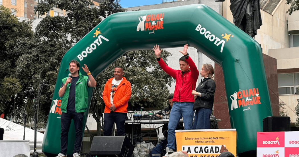 Director del Instituto de Bienestar Animal de Bogotá sobre una tarima bajo un arco inflable verde con logos de Bogotá y “Guau al Parque”, junto a equipos de sonido y un cartel informativo, durante una actividad pública al aire libre en una plaza de Bogotá.