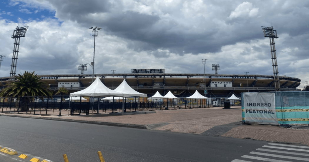 Vista exterior de una terminal de transporte con varias carpas blancas alineadas en la zona de acceso. Al fondo se observa un edificio de varios niveles y un cielo nublado con abundantes nubes grises. En primer plano hay una amplia superficie pavimentada sin presencia visible de personas.