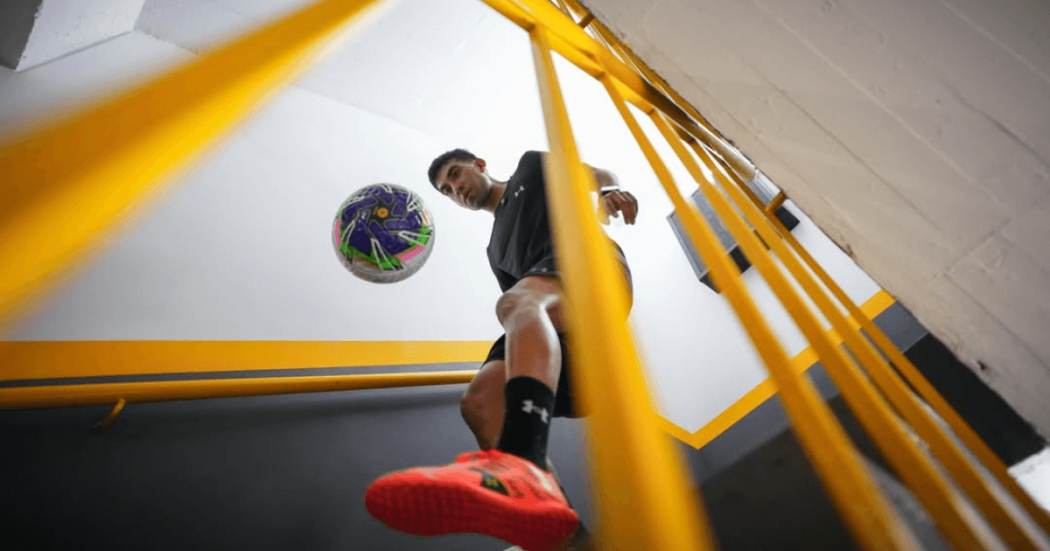 Messedinho realizando dominio de balón de fútbol en una escalera interior, vista desde abajo entre barandas amarillas que enmarcan la escena.