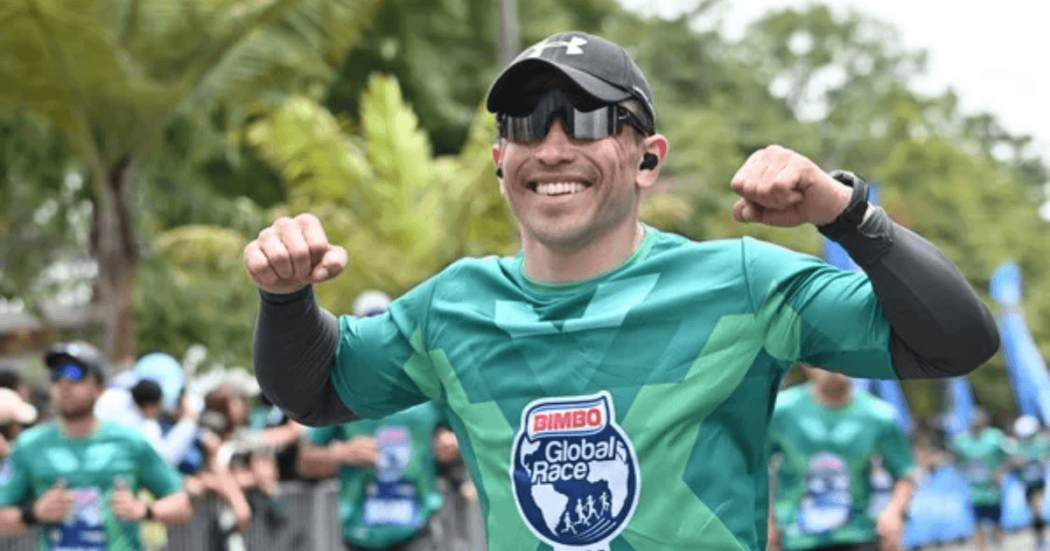 Participante de una carrera atlética viste camiseta verde de la Bimbo Global Race, gorra negra, gafas oscuras y audífonos inalámbricos, mientras avanza por el recorrido junto a otros corredores en un entorno al aire libre rodeado de árboles.