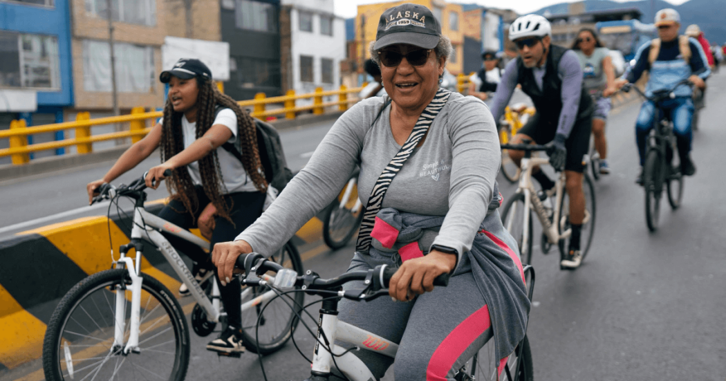 Personas en bicicleta recorren una vía urbana durante una jornada de Ciclovía. En primer plano, una persona vestida con ropa deportiva gris y gorra conduce una bicicleta blanca