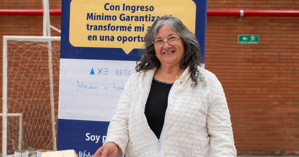 Ciudadana mayor posa en un stand de su emprendimiento en un evento organizado por la Secretaría Distrital de Integración Social, teniendo tras de ella un pendón sobre el programa Ingreso Mínimo Garantizado (IMG).