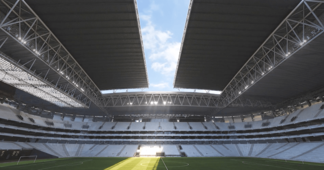 Vista interior del nuevo estadio El Campín de Bogotá con graderías, cancha de césped en el centro y una gran cubierta metálica parcialmente abierta que deja ver el cielo. La luz del sol ingresa por la apertura del techo e ilumina una franja del campo de juego.