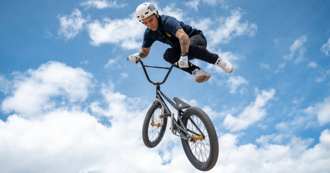 Luis Oswaldo Rincón ejecuta una maniobra aérea en una bicicleta BMX durante una competencia de ciclismo extremo. La bicicleta se encuentra inclinada en el aire y el deportista viste casco, camiseta oscura y pantalón claro.