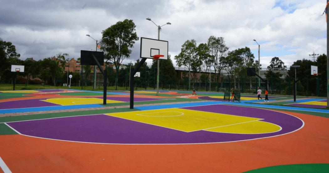 Cancha deportiva multicolor con tablero y aro de baloncesto, rodeada por cerramiento metálico, árboles y cielo nublado al fondo.