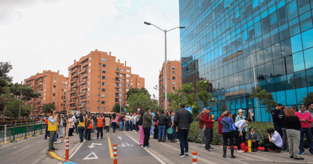 Personas reunidas en un espacio público junto a edificios de Bogotá durante una actividad de simulacro de evacuación. Varias personas permanecen organizadas sobre la vía, delimitada con conos de tránsito, mientras personal de apoyo coordina la jornada.