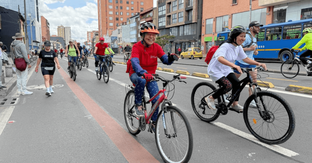 Ciclistas y peatones transitan por una vía urbana durante una jornada recreativa. En primer plano, dos personas montan bicicleta con casco mientras otras personas avanzan en bicicleta y caminando por la calzada.