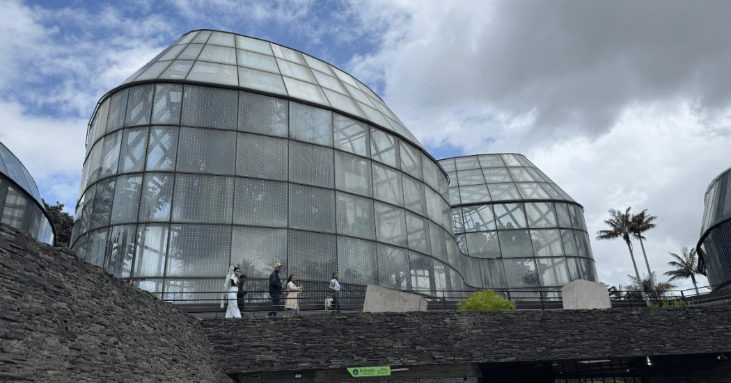 Vista exterior de los domos de vidrio del Tropicario del Jardín Botánico de Bogotá. En primer plano se observa un sendero elevado por el que transitan varias personas, mientras al fondo se aprecia un cielo parcialmente nublado y vegetación alrededor de la estructura.