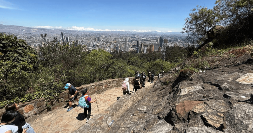 Personas recorren el sendero peatonal de Monserrate entre rocas y vegetación; al fondo se observa una vista panorámica de Bogotá bajo cielo despejado.