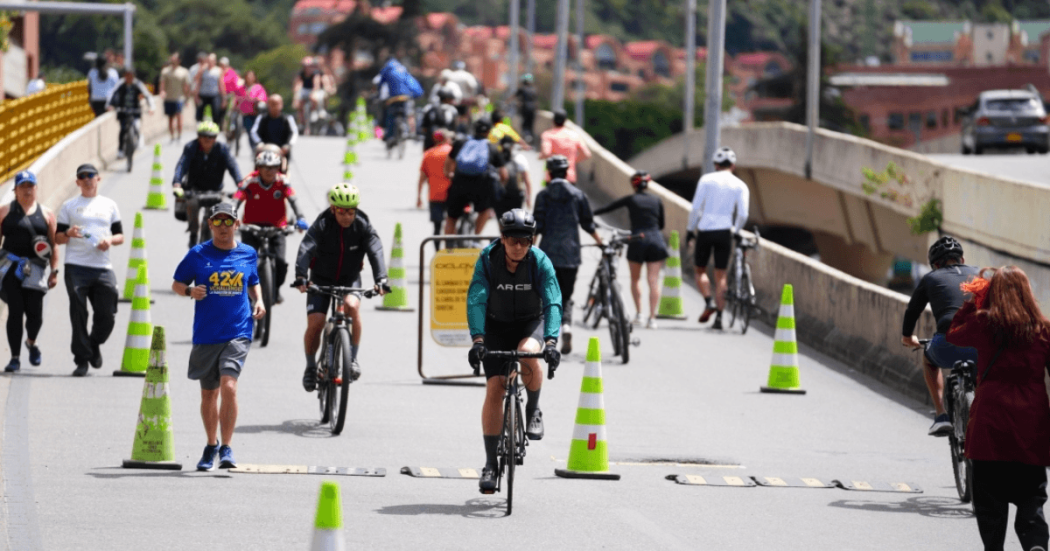 Personas caminando, trotando y montando bicicleta por una vía habilitada para la Ciclovía de Bogotá, delimitada con conos verdes y blancos, sobre un puente urbano durante el día.