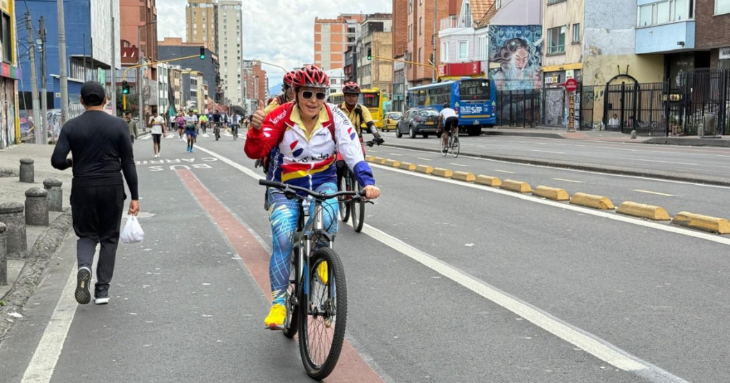 Persona con camiseta blanca, roja y azul monta una bicicleta sobre una vía de la Ciclovía de Bogotá; al fondo se observan edificaciones, vehículos y otros ciclistas.