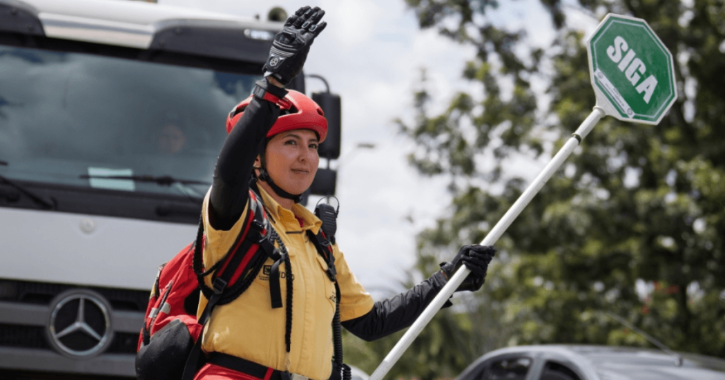 Persona con casco rojo, guantes y uniforme amarillo sostiene una paleta de señalización con la palabra “SIGA” mientras levanta una mano.