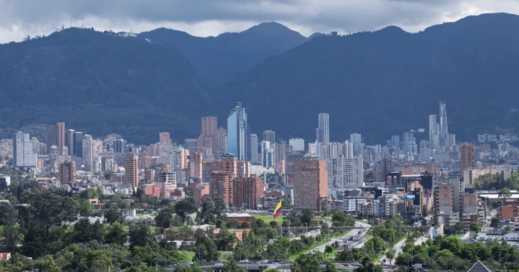 Panorama de Bogotá con vista amplia de edificaciones y rascacielos en el centro de la ciudad, zonas verdes en primer plano y los Cerros Orientales al fondo bajo un cielo parcialmente nublado.