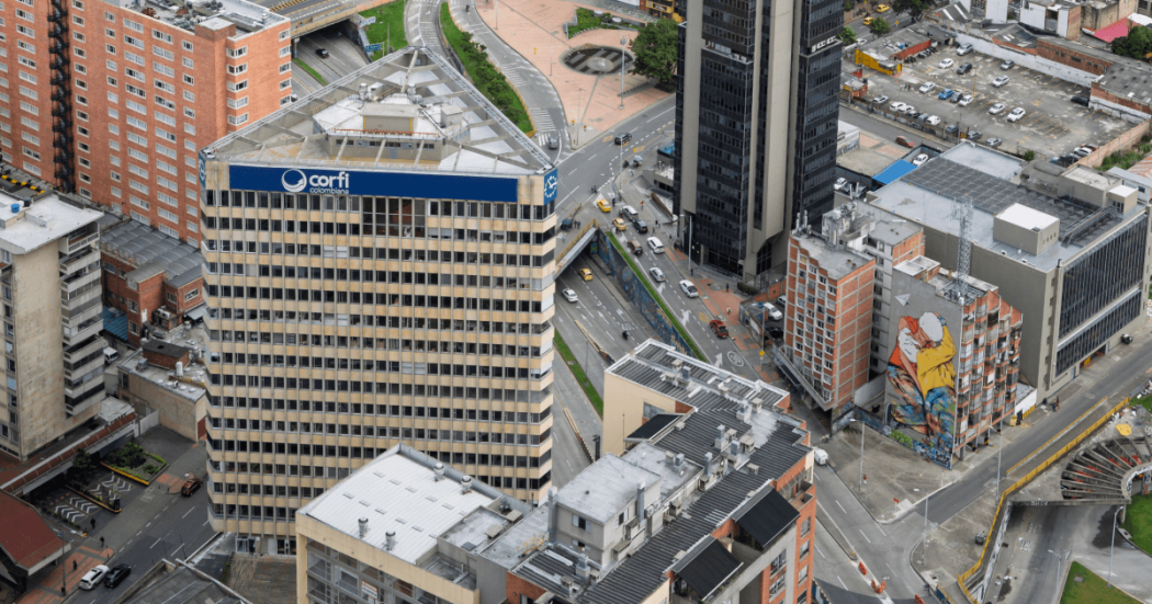Vista aérea de edificios y vías en el centro de Bogotá. En primer plano destaca un edificio con el letrero “Corfi Colombiana”, rodeado de torres de oficinas, calles con vehículos en circulación y un mural de gran formato en la fachada de un edificio cercano.