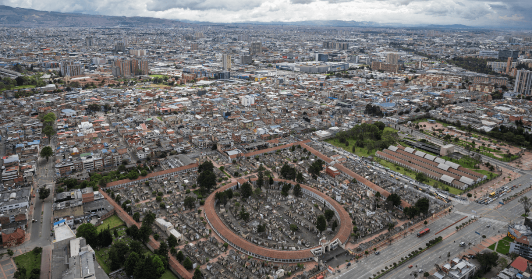 Vista panorámica de Bogotá con edificaciones residenciales y comerciales, corredor vial rodeado de árboles, amplias zonas verdes, cementerio y conjuntos de apartamentos al fondo bajo cielo nublado.