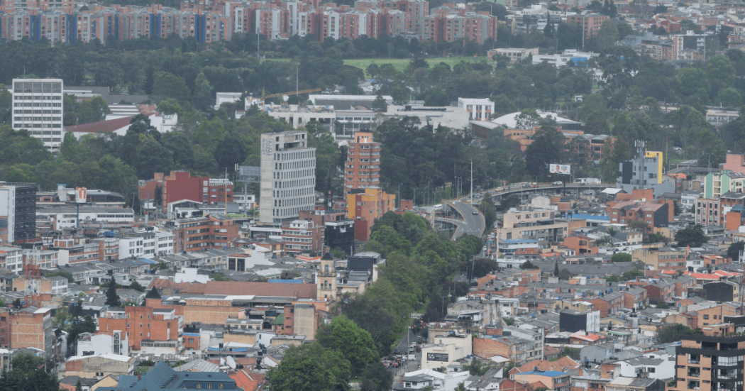 Vista panorámica de Bogotá con edificaciones residenciales y comerciales, corredor vial rodeado de árboles, amplias zonas verdes y conjuntos de apartamentos al fondo bajo cielo nublado.