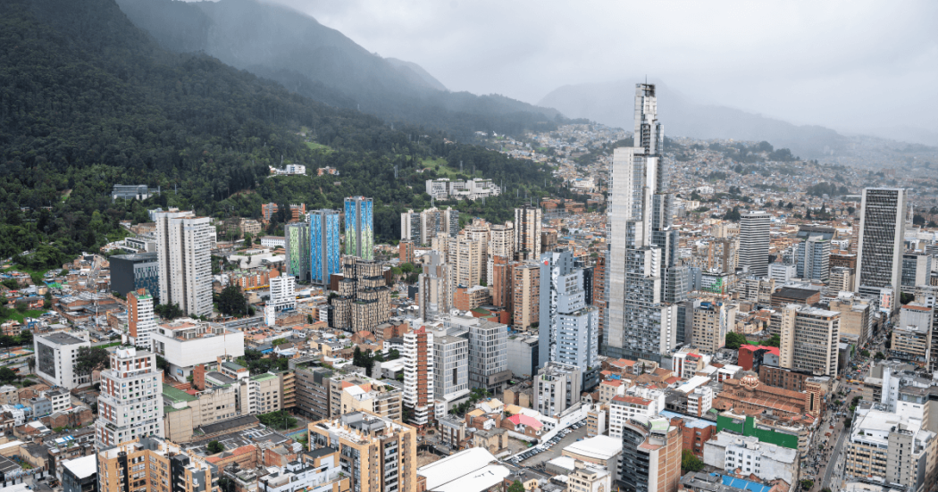 Vista panorámica de Bogotá con edificios de gran altura y zonas residenciales, junto a los cerros cubiertos de vegetación y neblina al fondo bajo un cielo nublado.