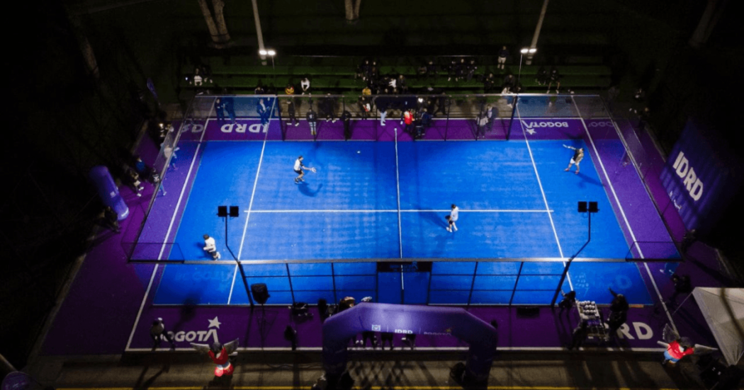 Vista aérea de varias canchas de pádel iluminadas en color azul dentro de un complejo deportivo cubierto, con jugadores en acción y áreas de circulación alrededor de los escenarios deportivos.