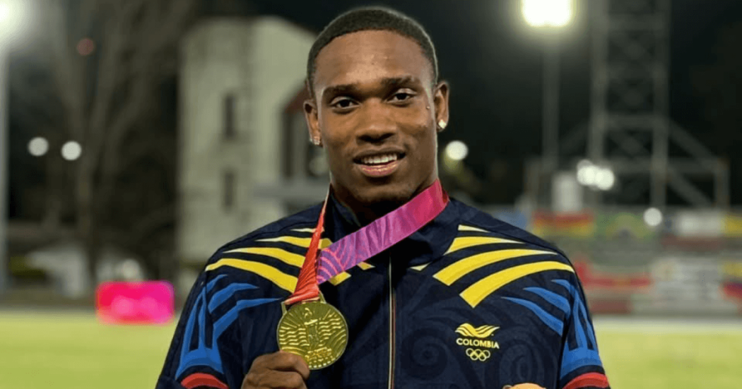 Ronal Longa sostiene una medalla dorada frente a una pista de atletismo iluminada durante la noche. En la prenda se observa el logo del Comité Olímpico Colombiano.