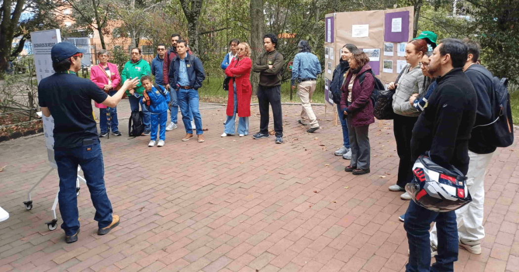 Personas participan en una actividad al aire libre sobre una plazoleta de ladrillo. Varias están alineadas y observan una demostración; algunas llevan chaquetas azules y una persona en el centro viste una chaqueta roja.